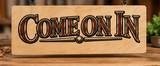 Handcrafted wooden Come On In welcome sign featuring bold vintage-style engraved lettering and a natural wood finish, displayed in a warm rustic workshop.